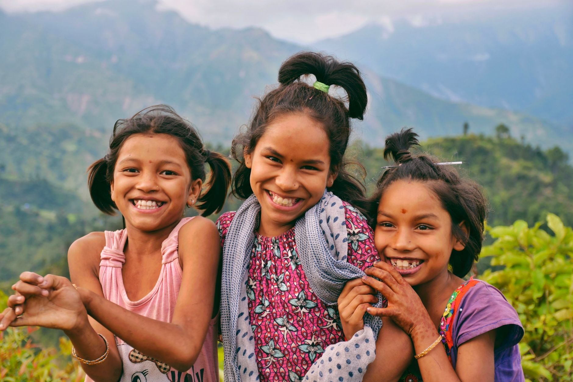 Three joyful South Asian children smiling outdoors in the lush mountains of Nepal, exuding happiness and natural beauty.